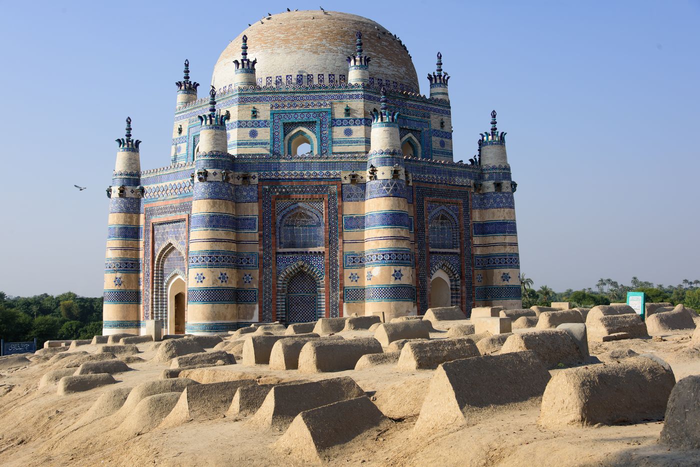 An octagonal tomb faced with blue and white glazed tiles, with corner turrets and a broad dome, rising behind rows of mud-plastered graves under a clear sky.