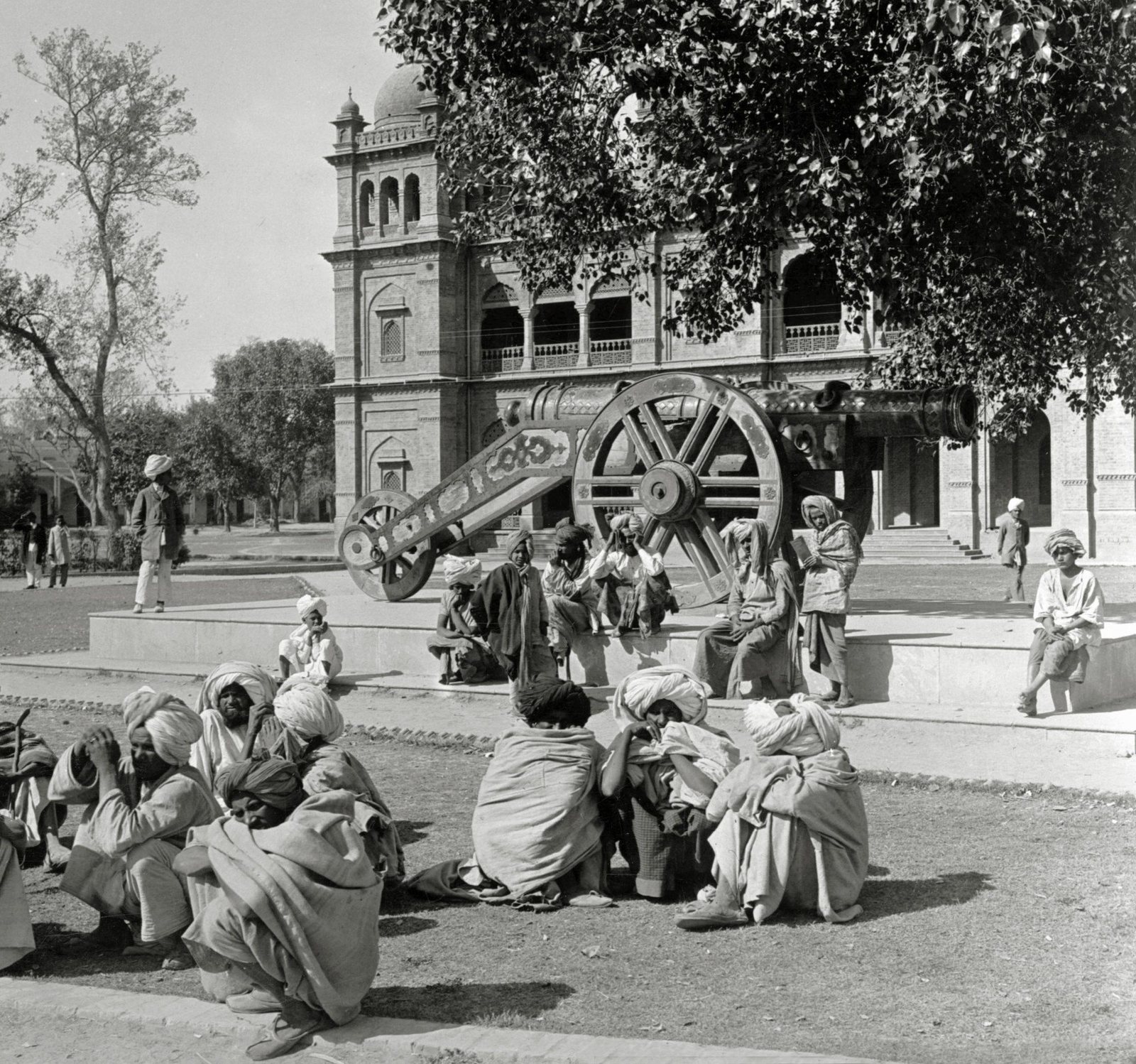 Old photograph of a huge bronze cannon on a wheeled carriage, set on a raised platform in front of a domed museum building, with men in turbans and shawls sitting on the platform and on the grass.